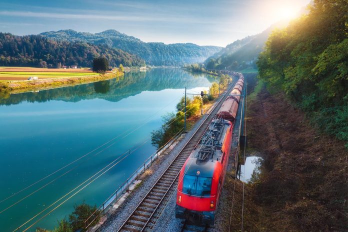 Aerial view of red train moving near river in alpine mountains