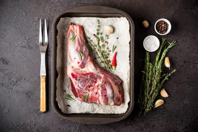 Raw fresh Lamb Meat in baking tray, herbs and fork on black stone background.