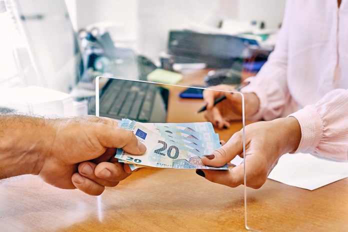 Hand of female social worker taking or giving euro money from client at office workplace with plexiglass separator.